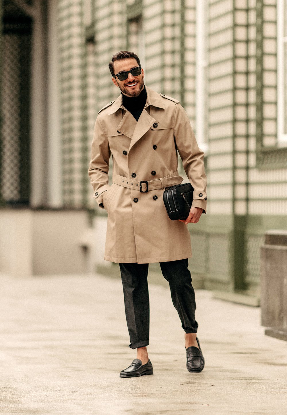 Beige trenchcoat med dubbelknappning och bälte. Svart polokrage, svarta byxor och svarta loafers. Liten svart väska i handen.
