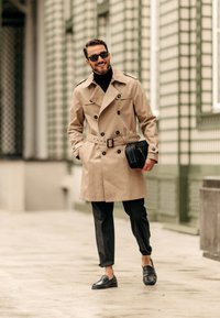 Beige trenchcoat med dubbelknappning och bälte. Svart polokrage, svarta byxor och svarta loafers. Liten svart väska i handen.