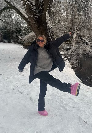 Chaqueta acolchada negra, suéter gris, jeans negros y botas de invierno rosas sobre tierra nevada, con árboles al fondo.