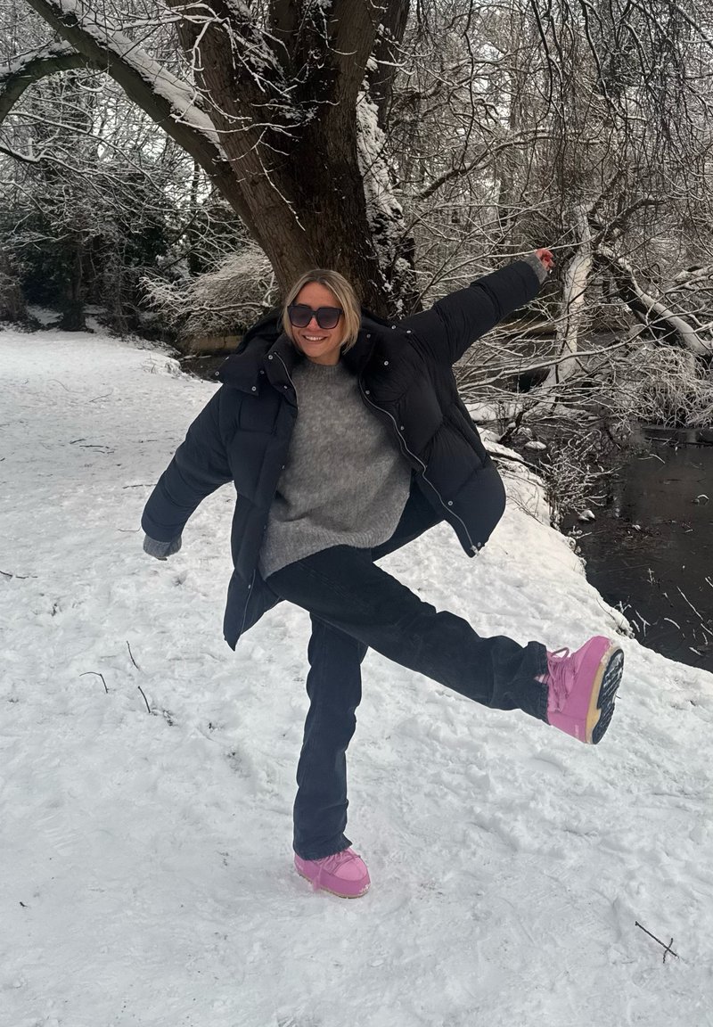 Doudoune noire, pull gris, jean noir et bottes d'hiver roses sur un sol enneigé, avec des arbres en arrière-plan.