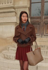 Bruine wolblendjas met dubbel knoopsgat, grijze sjaal, bordeauxrode geplooide rok, zwarte leren handschoenen en beige tas met oranje voering.