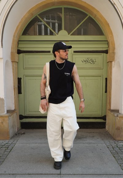 Camisa negra sin mangas, pantalones blancos holgados, gorra negra, gafas de sol y zapatos negros. De pie frente a una puerta verde con detalles decorativos.