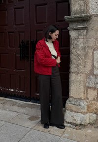 Γυναίκα με κόκκινο μπουφάν και μαύρο φαρδύ παντελόνι στέκεται δίπλα σε μεγάλη, σκοτεινή ξύλινη πόρτα και τοίχο από πέτρα, κοιτάζοντας προς τα κάτω και ρυθμίζοντας το μανίκι του μπουφάν της.