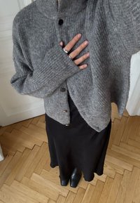 Cardigan gris en maille avec un col haut et de gros boutons, associé à une robe noire et des bottines noires épaisses. Fond en parquet en bois.