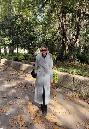 Mujer con un abrigo largo gris con cuello de piel, gafas de sol y pantalones negros caminando por una acera cubierta de hojas junto a áreas verdes y árboles.