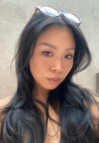 Jeune femme aux longs cheveux foncés et à la peau claire, portant des lunettes écaille de tortue sur la tête, devant un mur clair carrelé.