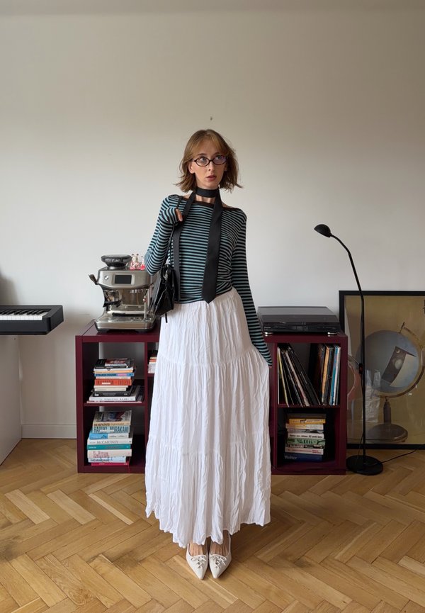 Jeune femme avec des lunettes, un chemisier à manches longues rayé, une écharpe noire, une jupe blanche et des chaussures pointues, debout dans une pièce moderne avec des étagères et un clavier.