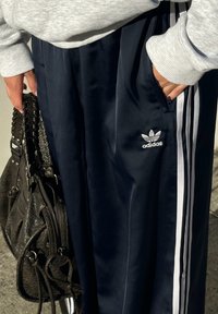 Calças desportivas azul-marinho feitas de material brilhante, apresentando riscas laterais brancas e um logótipo da Adidas bordado na coxa esquerda.