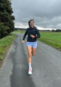 Runner in a black zip-up jacket and light blue shorts, wearing white and pink running shoes, on a cloudy road lined with greenery.