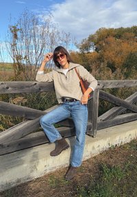 Bež, teksturiran pulover z zadrgo, modri ravni hlače in rjavi gležnjarji. Lesena ograja in jesensko listje v ozadju.