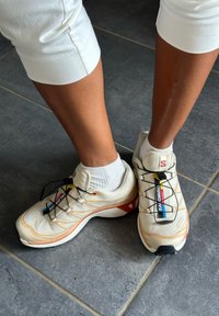 Jasno beżowe buty sportowe z teksturowaną siateczką, czarnymi sznurówkami oraz czerwonymi i niebieskimi akcentami, noszone z białymi skarpetkami do kostek.