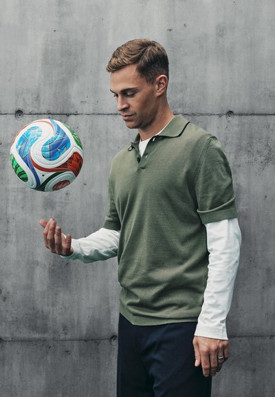 Camisa polo verde de manga corta sobre una camisa blanca de manga larga, sosteniendo un balón de fútbol colorido contra un fondo de concreto gris.