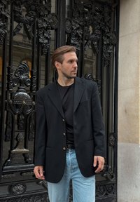 Black blazer with gold buttons, worn over a black shirt, paired with light blue jeans. The background features ornate black iron gates.