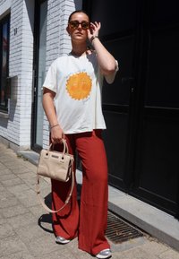 Χαλαρή κρέμα μπλούζα με γραφικό ήλιο, σε συνδυασμό με φαρδιά κόκκινα παντελόνια, μπεζ τσάντα και ασημένια sneakers. Ζεστή εξωτερική ατμόσφαιρα.