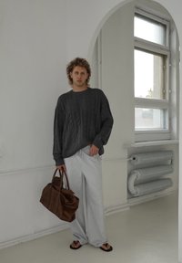 Suéter cinza de malha canelada, calças largas listradas claras, bolsa de couro marrom e chinelos, ambientado em um interior minimalista com janelas.