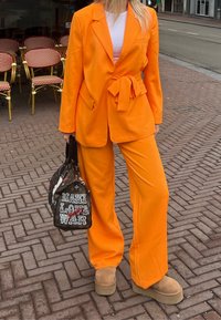 Blazer orange ajusté avec un pantalon assorti, présentant une ceinture à la taille. Chaussures à plateforme marron et un sac noir à motifs avec texte.