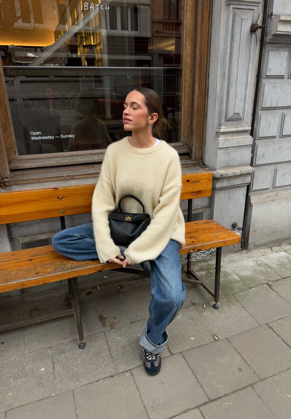 Maglione oversize crema, jeans in denim blu, borsa nera, seduta su una panchina di legno. Ambientazione all'aperto con una vetrina di negozio sullo sfondo.