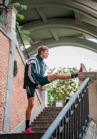 Casaco atlético preto com riscas brancas, calções pretos e ténis de corrida cor-de-rosa brilhante. Indivíduo a alongar-se em degraus de pedra sob um arco.