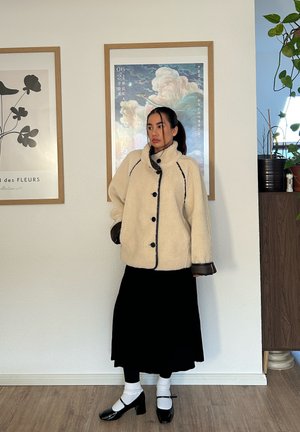 Chaqueta esponjosa de color crema con ribete y botones negros, combinada con una falda negra, calcetines blancos y zapatos negros. Fondo interior.