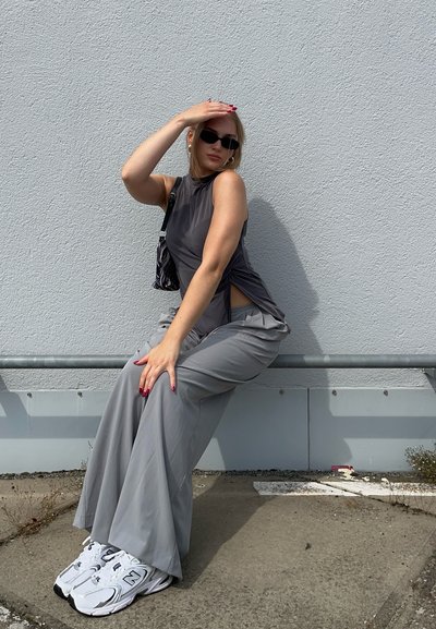Top gris sin mangas con aberturas laterales, pantalones gris de pierna ancha y zapatillas blancas. Un pequeño bolso a rayas descansa a un lado. Texturas lisas visibles.
