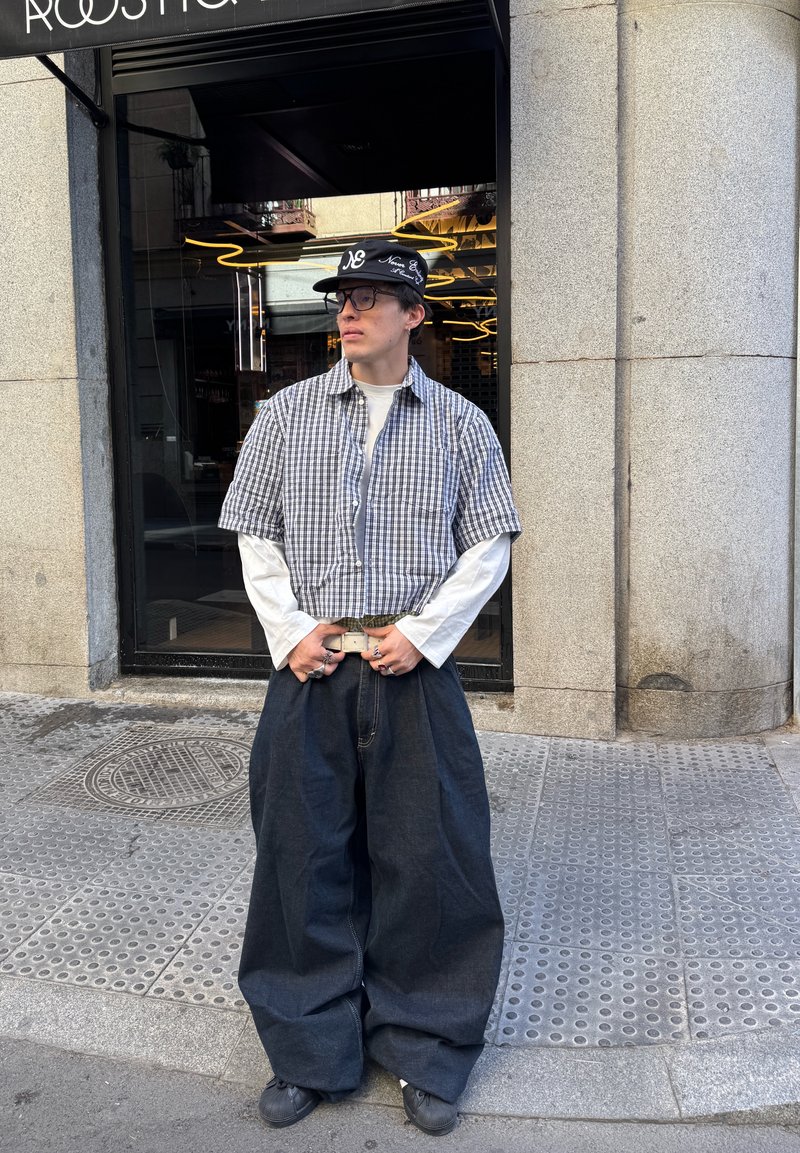 Chemise à manches courtes à carreaux noir et blanc sur une chemise à manches longues blanche ; jean ample en denim foncé ; casquette noire avec des accents blancs ; ceinture en argent.