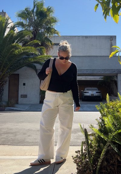 Top negro de manga larga, pantalones anchos blancos con textura, sandalias claras con rayas negras y bolsa de mimbre. Palmeras y un coche al fondo.