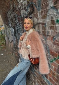 Giacca in pelliccia finta rosa, indossata sopra una leggera blusa in satin con dettagli in pizzo, abbinata a jeans a gamba larga con un pannello scuro. Inclusa una borsa marrone.