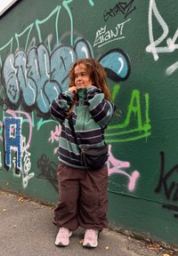 Otrok oblečen v zeleni in črni prugasti flis, rjave cargo hlače in roza superge, z črnim pasom, ob graffiti steni.