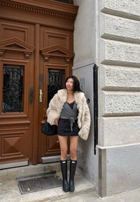 Ung kvinde i beige, pelsagtig jakke, grå top, sort mininederdel og sorte gummistøvler står ved en brun, udsmykket dobbeltdør på en brolagt gade.