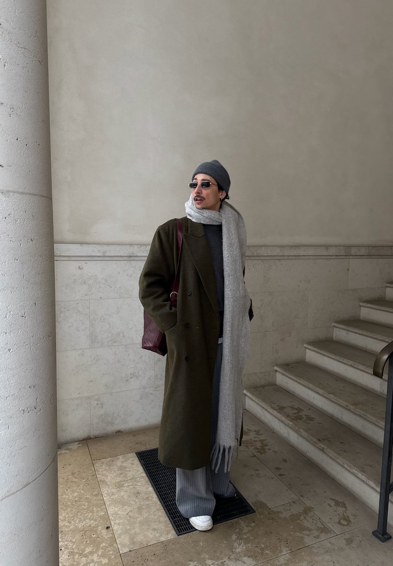 Homme portant un long manteau foncé, une écharpe grise, un bonnet, des lunettes de soleil et des chaussures blanches, debout près d'un escalier en pierre et d'une grande colonne à l'intérieur.