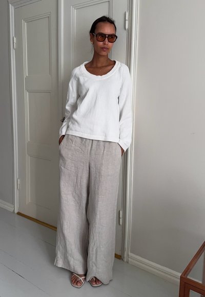 Mujer con gafas de sol marrones, suéter de punto blanco, pantalones de lino beige de pierna ancha y sandalias blancas, de pie en el interior junto a una puerta cerrada.