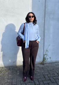 Blusa de manga longa azul clara com decote franzido, combinada com calças largas castanhas e sapatos de bico fino vermelhos escuros. Bolsa castanha incluída.