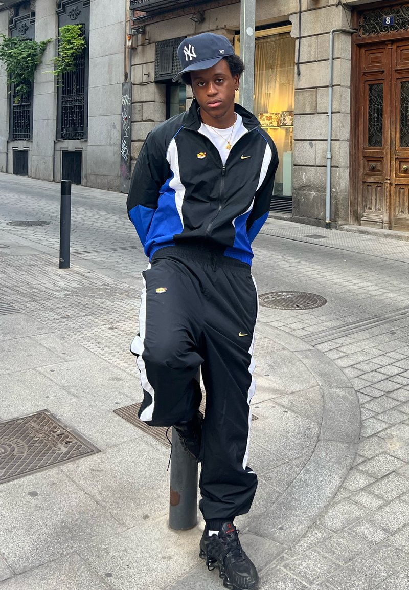 Survêtement noir et bleu avec des accents blancs et le logo Nike, associé à des baskets noires. Debout devant un décor urbain.