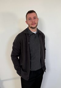 Νεαρός άνδρας με κοντά μαλλιά και γένια, φοράει σκούρα ζακέτα πάνω από γκρι πόλο πουκάμισο, στέκεται μπροστά σε μια απλή λευκή τοίχο με τα χέρια στις τσέπες.