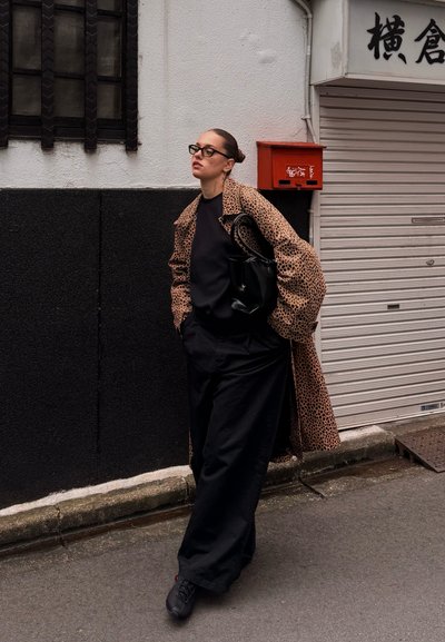 Abrigo trench con estampado de leopardo sobre un suéter negro, combinado con pantalones negros de pierna ancha y un bolso negro estructurado. Ambiente urbano casual.