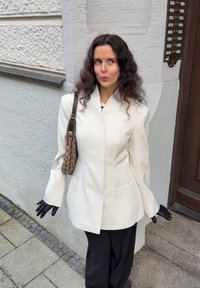 White tailored blazer with wide sleeves and pockets, worn over a striped top. Black gloves and a brown patterned shoulder bag complete the look.