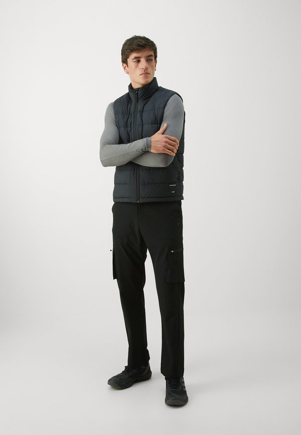 Czarna puchowa kamizelka z wysokim kołnierzem, noszona na jasnoszarym, długim rękawie. Czarne spodnie cargo z bocznymi kieszeniami i ciemne buty.