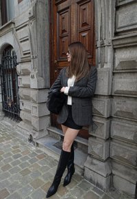 Szürke kockás blézer, fehér pulóver, fekete miniszoknya és térdig érő fekete hegyes orrú csizma. Texturált kőfal és faajtó a háttérben.