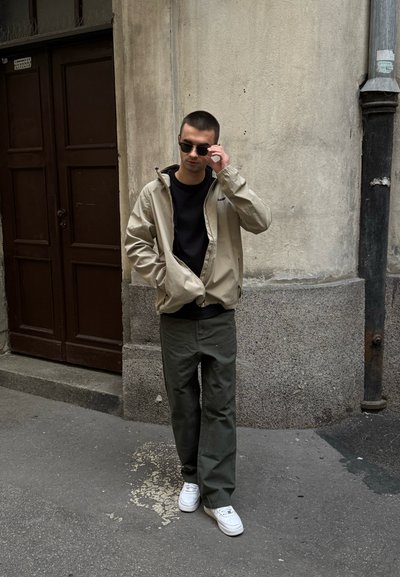 Chaqueta beige con cremallera sobre una camiseta negra, pantalones verde olivo y zapatillas blancas. Las gafas de sol añaden un detalle elegante contra un fondo texturizado.