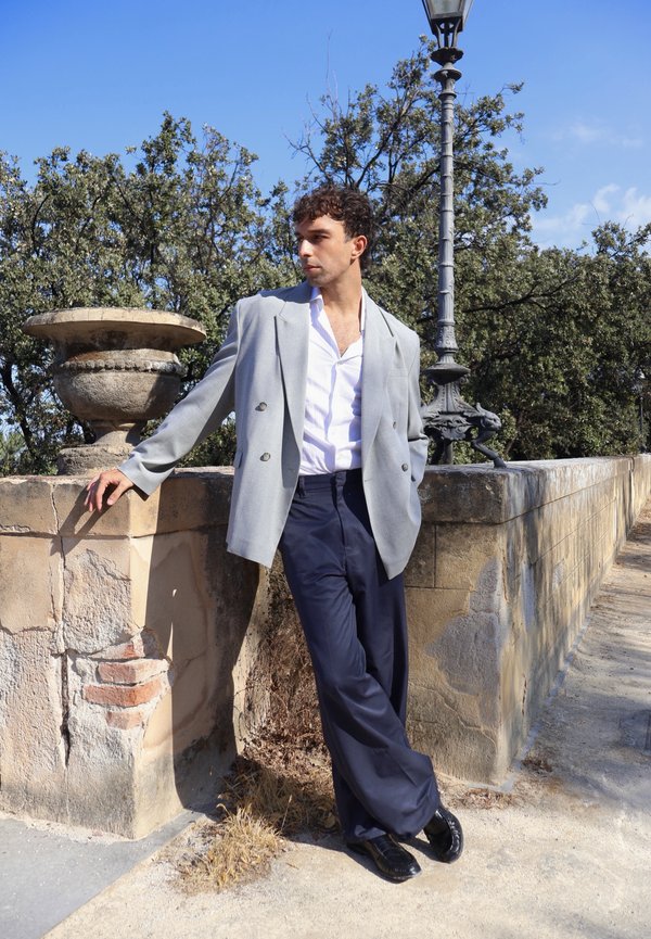 Homme en blazer gris et pantalon foncé appuyé contre un mur en pierre avec une urne et un lampadaire à proximité, regardant sur le côté par une journée ensoleillée.