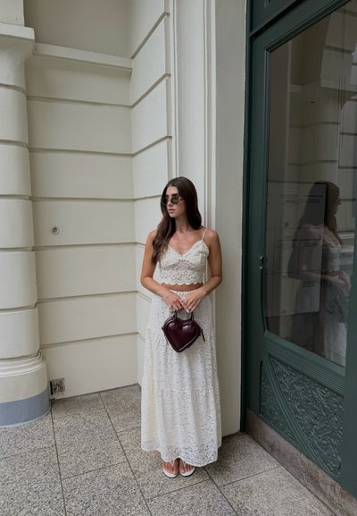 Top de encaje y falda larga blanca de encaje, combinados con un pequeño bolso burdeos. De pie cerca de una puerta verde, lleva gafas de sol redondas.