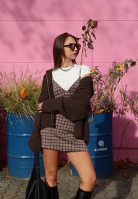 Smeđi oversized kardigan, bijela majica, karirana haljina tamnih boja, crne čizme do koljena i sunčane naočale na ružičastoj pozadini.