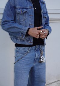 Light blue denim jacket and jeans, black polo shirt underneath, chain and keychain detail at the waist, multiple silver rings on hands.