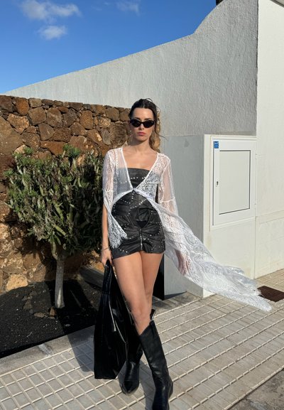 Cárdigan de encaje blanco sobre un vestido mini de cuero negro, con tachuelas plateadas y cremallera frontal, combinado con botas negras hasta la rodilla.