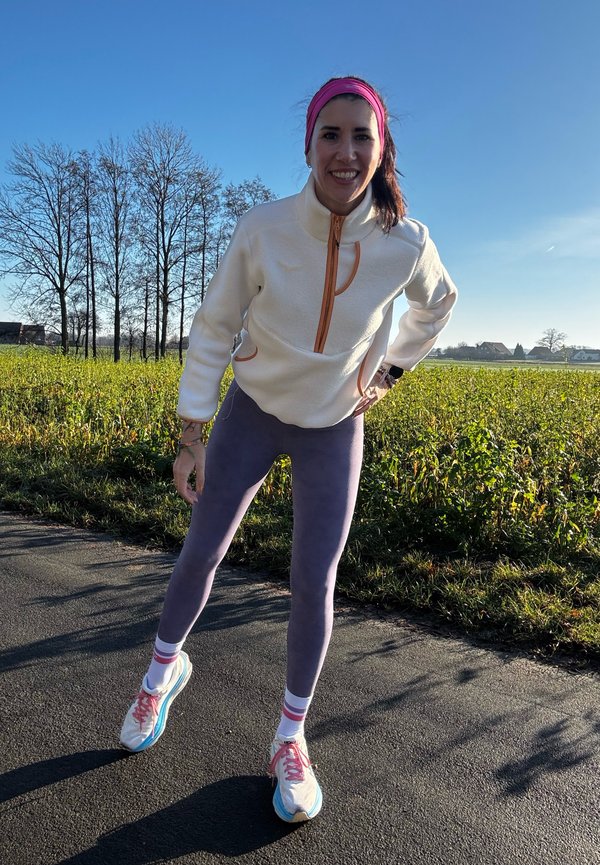 Fleece-Pullover in Weiß mit orangefarbenem Reißverschluss, kombiniert mit lila Leggings. Sportschuhe in Blau, Pink und Weiß mit gestreiften Socken.
