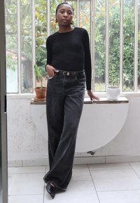 Mujer con una blusa negra de manga larga y jeans de pierna ancha está de pie en el interior junto a un alféizar de ventana con dos plantas en macetas, posando con confianza.