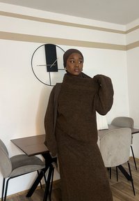 Ensemble marron tricoté en deux pièces avec un pull à col haut et une jupe ajustée. Texture douce, coupe décontractée et détails côtelés. Fond d'intérieur minimaliste.