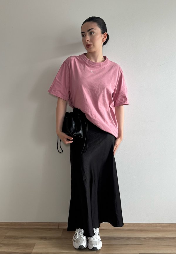 Donna con capelli scuri raccolti in uno chignon, che indossa una maglietta rosa oversize, una gonna lunga nera, sneakers bianche e tiene una piccola borsa nera davanti a un muro semplice.