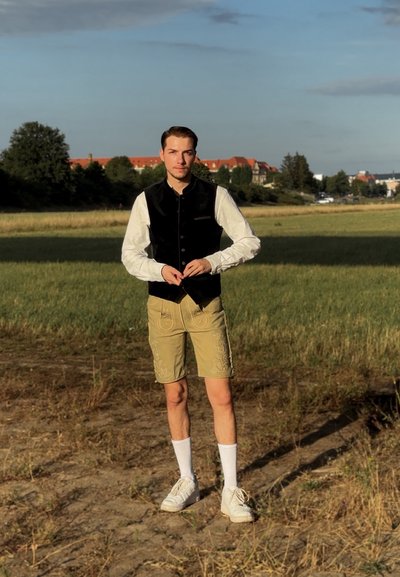 Chaleco de terciopelo negro sobre una camisa blanca de manga larga, combinado con pantalones cortos beige con bordado, calcetines blancos y zapatillas blancas.