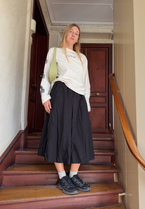 Jeune femme debout sur des escaliers en bois, portant une chemise blanche à manches longues, une jupe noire, des baskets noires, des chaussettes blanches et un sac à épaule vert clair.